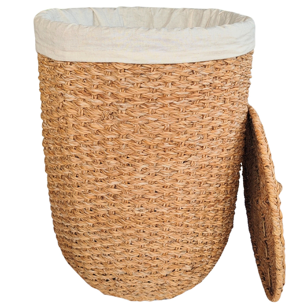 Cesto portabiancheria aperto in alghe marine di colore naturale, con inserto in tessuto chiaro e coperchio appoggiato lateralmente.