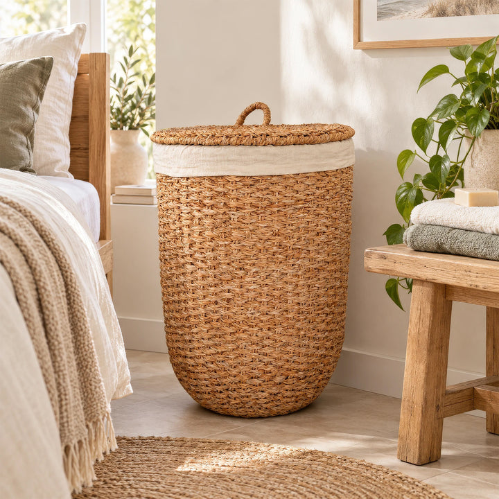 Cesto portabiancheria con coperchio, realizzato in alghe marine di colore naturale e con un inserto in tessuto chiaro, posizionato tra il letto e la panca in legno nella camera da letto.