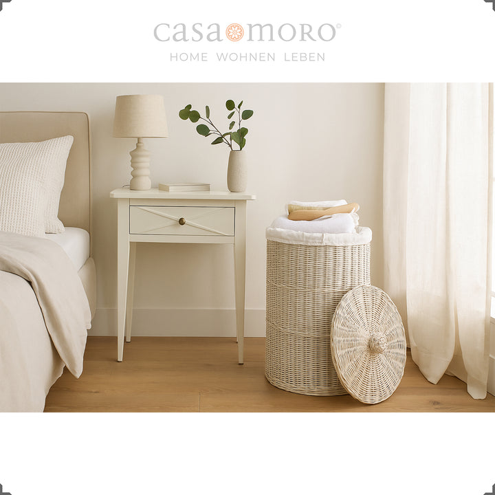 Weiß gekalkter Wäschekorb aus Rattan im Schlafzimmer, kombiniert mit hellen Textilien und natürlichem Licht